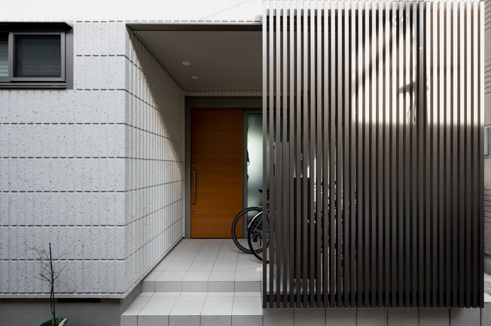 Modern house entrance featuring dark metallic slatted composite cladding privacy screen alongside textured white brick wall and wooden front door.