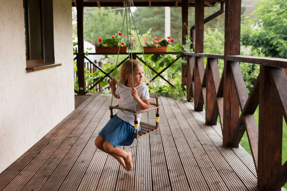 A young girl sitting on a suspended wooden swing on a covered patio featuring dark timber decking boards and a protective wooden cross-brace balustrade.